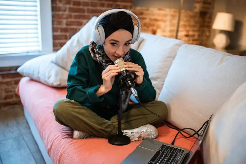 A young woman wearing headphones records a podcast at home using a microphone and laptop